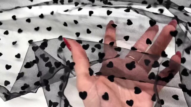 This delicate sheer tulle fabric features a charming black heart polka dot pattern on a fine mesh base. The lightweight and translucent material is perfect for romantic fashion overlays and decor.