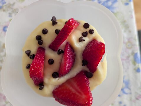 Dessert cremoso in coppa bianca, guarnito con fragole fresche e gocce di cioccolato, su tovaglia floreale dai colori delicati