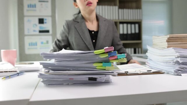 Overworked Asian businesswoman feeling stressed and tired with a large pile of documents on her desk. A corporate employee managing a heavy workload.