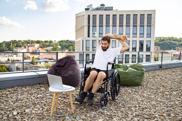 Obraz premium A man in a wheelchair stretches his arms on a rooftop terrace with a city view, beanbags, and a laptop nearby