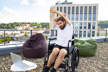 Obraz premium A man with a beard sits in a wheelchair on a rooftop terrace, stretching his arms while looking at a laptop on a small table