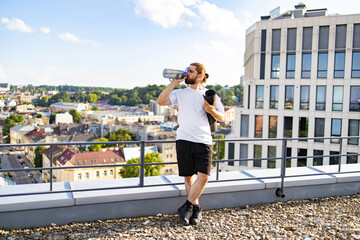 Obraz premium A man with long hair tied back drinks from a water bottle while holding a yoga mat on a rooftop overlooking a city
