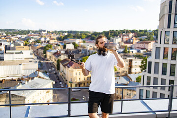 Obraz premium A man with headphones around his neck drinks from a metal water bottle while holding a selfie stick with a smartphone attached, overlooking a city skyline