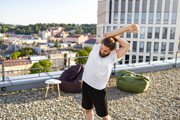 Obraz premium A man with a beard and bun hairstyle performs a shoulder stretch on a gravel rooftop with a city view and modern buildings in the background