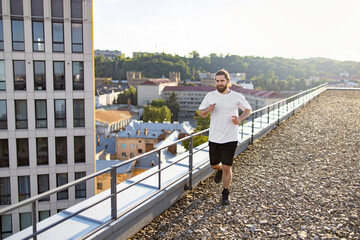 Obraz premium A bearded man in athletic wear jogs along a gravel rooftop with a railing, overlooking urban buildings and green hills under a bright sky