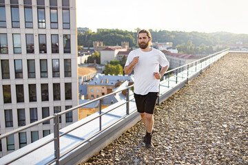 Obraz premium A fit man with a beard and long hair runs on a gravel rooftop terrace with a modern building and cityscape in the background during golden hour