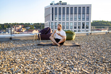 Obraz premium A man with a beard sits cross-legged on a yoga mat on a gravel rooftop terrace, eating from a bowl while beanbag chairs and a modern building are visible in the background