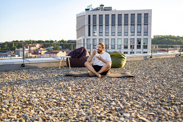 Obraz premium A man with a beard sits cross-legged on a yoga mat on a gravel rooftop, eating from a bowl with a fork, with beanbag chairs and a modern building in the background