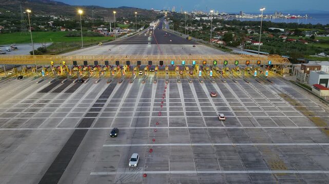 Wide aerial view of illuminated toll plaza at dusk with vehicles approaching highway lanes, drone