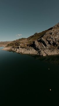 A vertical cinematic FPV drone shot flying over the Porma reservoir in Leon, Spain, featuring rocky shorelines, clear water reflections, and mountain landscapes.