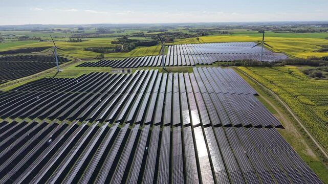 A side tracking drone shot flying over endless rows of solar panels combined with wind turbines, situated next to bright yellow rapeseed fields in the rural Danish countryside.