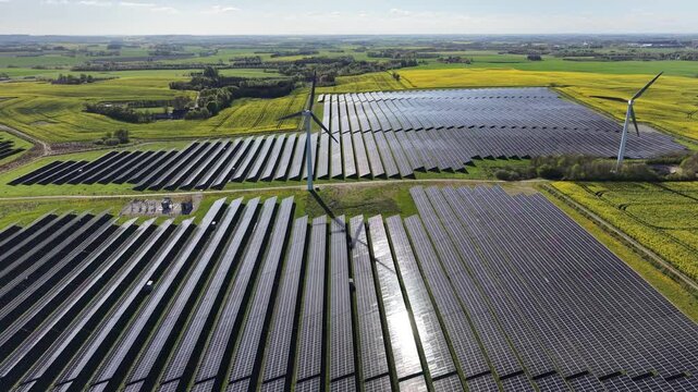 An orbiting drone shot moving smoothly over a large-scale solar installation integrated with wind power and into the agricultural fields of the Danish countryside.