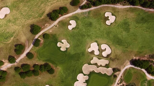 Top-down view of a golf course featuring multiple sand bunkers, manicured green fairways, winding cart paths and scattered trees across the landscape