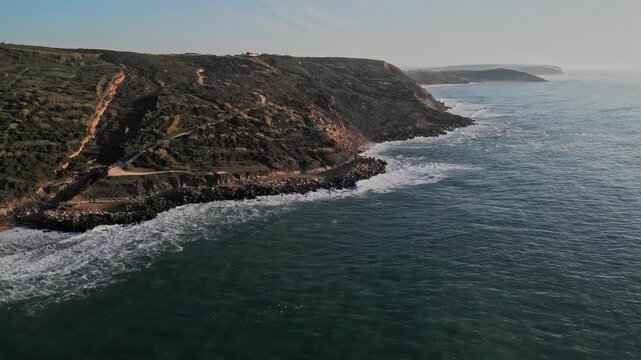 Cinematic aerial shot of the rugged coastline at Praia do Salgado in Nazar&eacute;, Portugal. The drone footage shows waves crashing against rocky cliffs under a clear sky