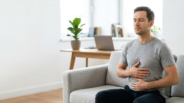 Man practicing deep breathing or meditation at home. Male experiencing stomach pain or discomfort in a modern living room. Copy space for text