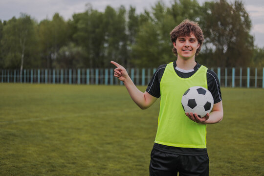 Young soccer player holding football and pointing aside on outdoor field. Smiling athlete in training bib standing on green pitch with copy space for sports or fitness concept.