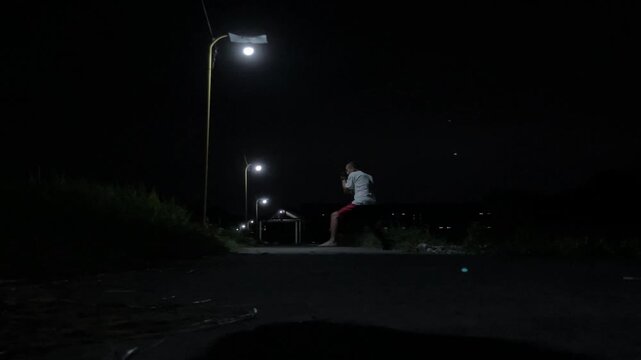 Cinematic 4K ground view of a man training Muay Thai shadowboxing under a street lamp in a dark, mysterious alley. Moody urban atmosphere and gritty fight practice.