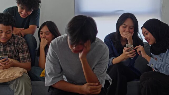 Distressed Asian man holding head in despair while group of friends laughs and uses phones behind