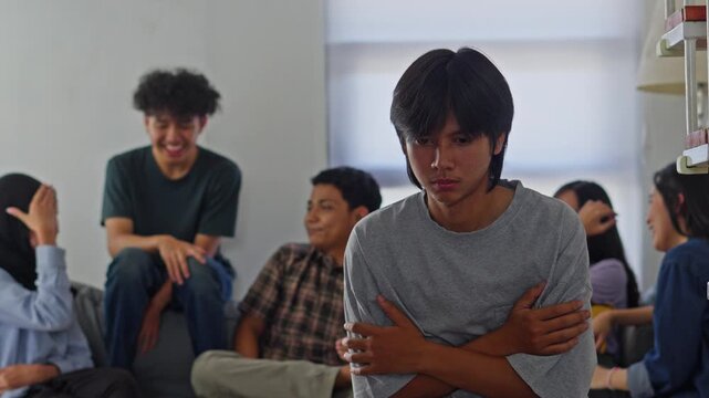 Introverted male youth looking downcast with arms crossed ignored by peers at gathering