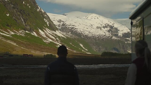 Couple steps outside their off grid camper, holding coffee and enjoying breathtaking glacier and mountain views in Norway. Cozy van life, freedom, and scenic outdoor lifestyle.