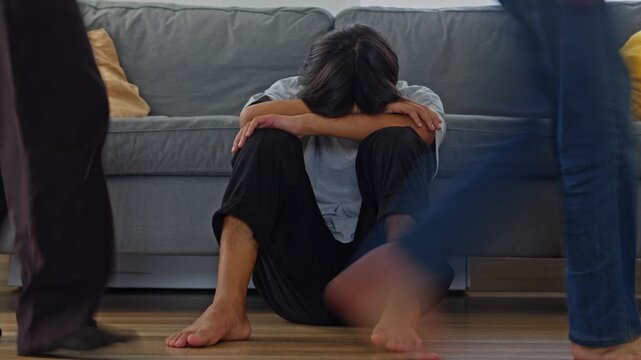 Asian teen hiding his face while sitting on the floor as blurred figures walk past