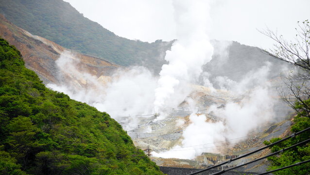地熱噴出孔から白い蒸気が立ち上る岩だらけの地形。緑の丘陵と工業施設が広がる山岳地帯の風景。