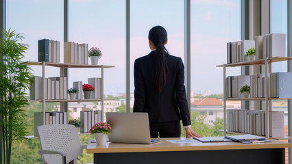 Professional woman looking out office window. ​Desk with laptop and pink flowers. ​White bookshelves filled with organized binders. ​Suburban view through large glass panels. ​Bright minimalist office © Jakub