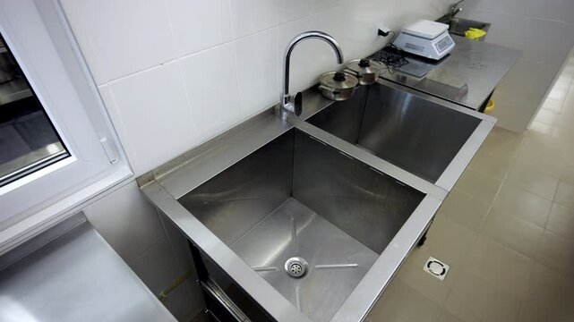 Double stainless steel sink with tall chrome faucet stands in professional kitchen. Digital scale and pots sit on adjacent metal table