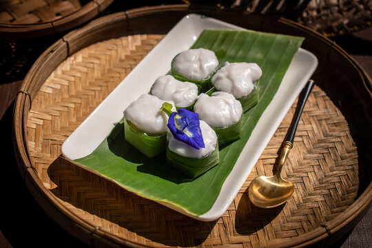 Traditional Thai pudding with creamy coconut topping in banana leaf cups, decorated with pea flower. Set on a wooden tray under natural sunlight with shadows. A delicious tropical dessert concept.