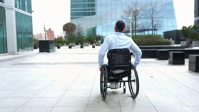 Back view of a young disabled businessman riding his wheelchair through a modern city financial district with glass buildings