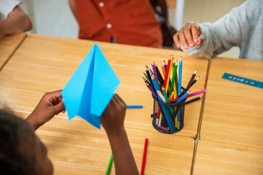 High angle view of elementary school student holding a blue paper airplane at a wooden desk with colored pencils. Creativity and imagination concept.
