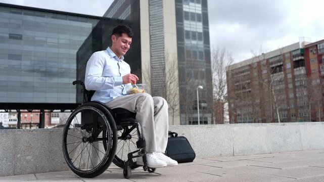 Young businessman with a physical disability eating a healthy salad for lunch. He is sitting in a wheelchair outside a modern office building