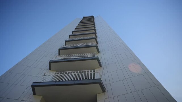 Tall modern residential building reaches toward clear blue sky. White facade panels and uniform balconies create geometric pattern