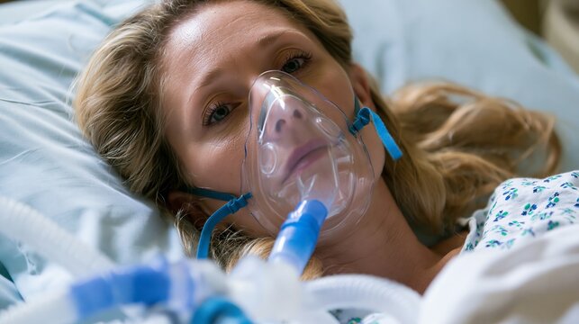 Woman in hospital bed, recovering from surgery. She wears an oxygen mask connected to breathing equipment, possibly for a coma or heart condition.