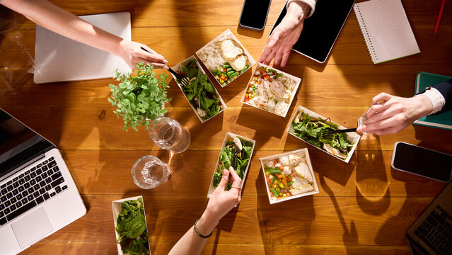 Top view of healthy office lunches with coworkers eating at wooden table. Concept of corporate catering, office meal plans, team lunch breaks and food delivery service promotion.