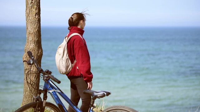 Young woman with brown hair in red jacket standing near bicycle by tree overlooking calm sea