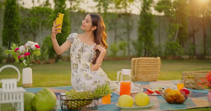Indian young adult gen z relax female use mobile make funny pout face on video call enjoy picnic trip. Teen girl hold phone look cam do pose take selfie photo have fun joy day spend time garden place