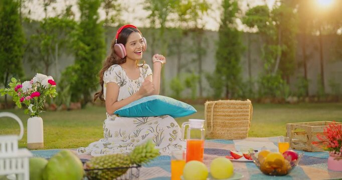 Indian gen z female sitting on mat wear headset listen good mp3 music sing song enjoy day fresh air garden Happy young adult relax girl dancing spend time feel free have fun joy at picnic trip place