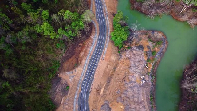 Cinematic 4K drone flight following a modern highway snaking through lush Thai rainforest. The camera pans up to reveal a vibrant green waterway and dense forest canopy at the border regions.