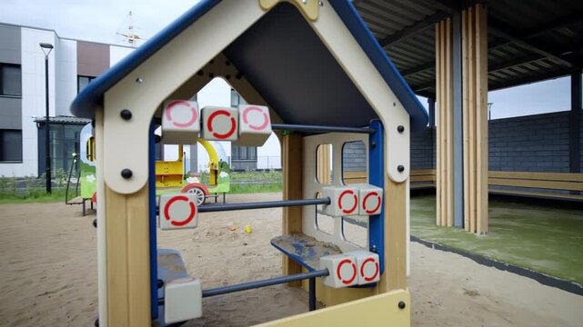 Small toy house with abacus stands on sandy playground near shelter. Brick wall and black door create industrial backdrop for play
