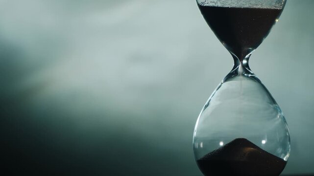 Close up of hourglass measuring passage of time with flowing sand against dark smoky background.