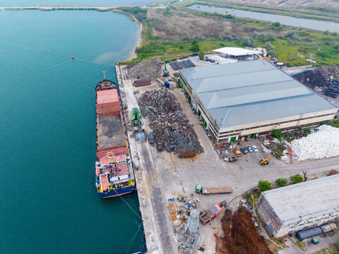 Cargo ship sits at dock near warehouse while recycling materials are piled on shore during daytime in industrial area by water