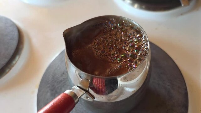 Coffee brewing in a metal pot on a stove. Foam bubbling as the drink reaches a boil. Traditional process of making hot dark caffeinated beverage for breakfast.