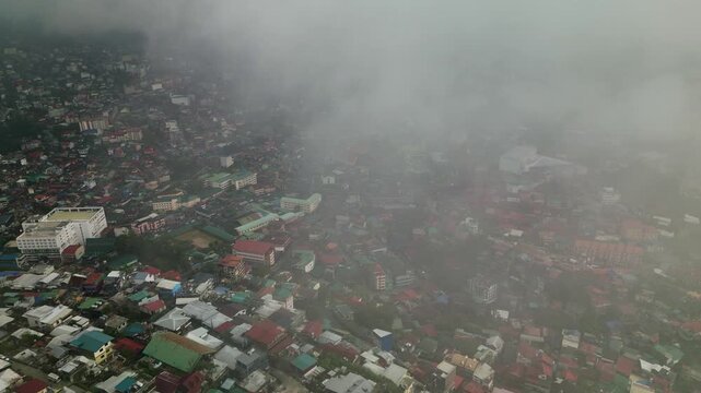 Aerial orbit of foggy urban landscape in Baguio City, Benguet, Philippines with dense buildings and low clouds covering the cityscape.