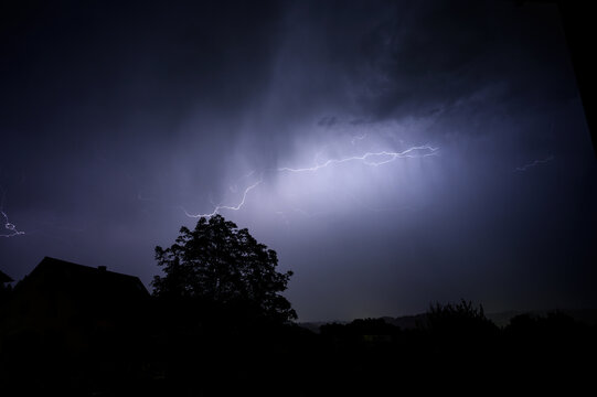 Dramatic lightning strikes illuminate the night sky over silhouetted trees and a house