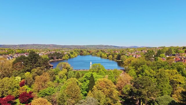 Aerial drone footage of Roath Park in Cardiff, Wales, capturing blue waters of the lake, lakeside greenery and the surrounding residential suburbs under a clear, blue sky. Ascending shot.