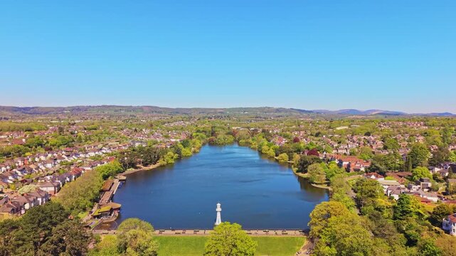 Aerial drone footage of Roath Park in Cardiff, Wales, capturing blue waters of the lake, lakeside greenery and the surrounding residential suburbs under a clear, blue sky. High altitude push-in shot.