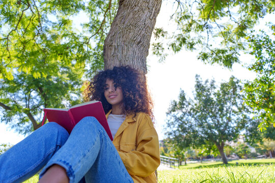 Young woman enjoying nature, reading book, sitting under tree in park, finding inspiration