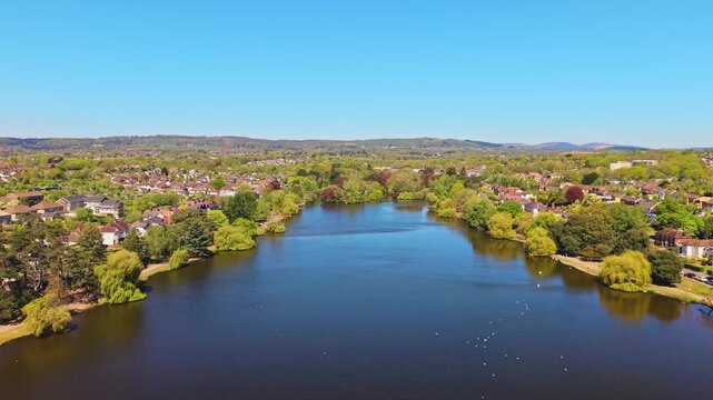 Aerial drone footage of Roath Park in Cardiff, Wales, capturing blue waters of the lake, lakeside greenery and the surrounding residential suburbs under a clear, blue sky. Pull-out shot.