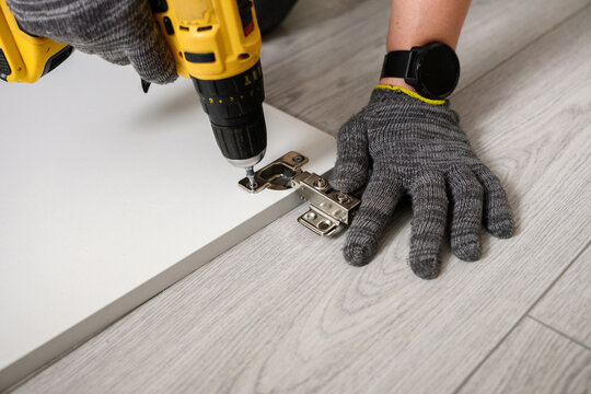 Professional furniture assembly using cordless drill driver. Skilled handyman installing cabinet hinges on white board. Expert power tool woodworking and furniture installation service concept.